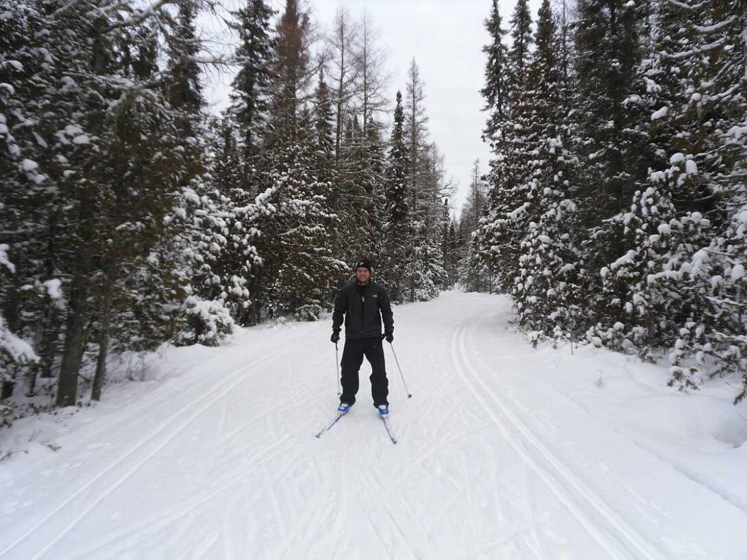 a cross-country skiier smiles at the camera as he skis down a snowy forest trail on a cloudy day.