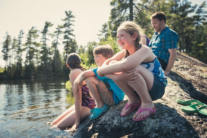 Some smiling children and a man sit on rocks next a lake on a sunny summer day. One child has their legs in the water and there are pine trees in the background.