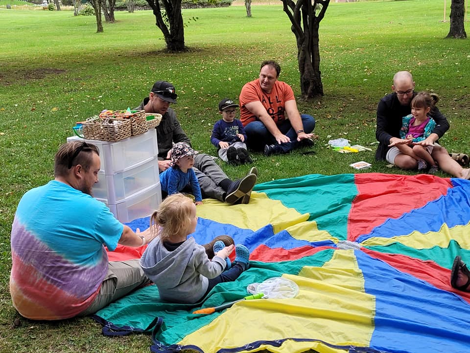 A group of parents and small children sit on a brightly coloured parachute in a green grassy park, smiling at each other. They are surrounded by toys and crafts.