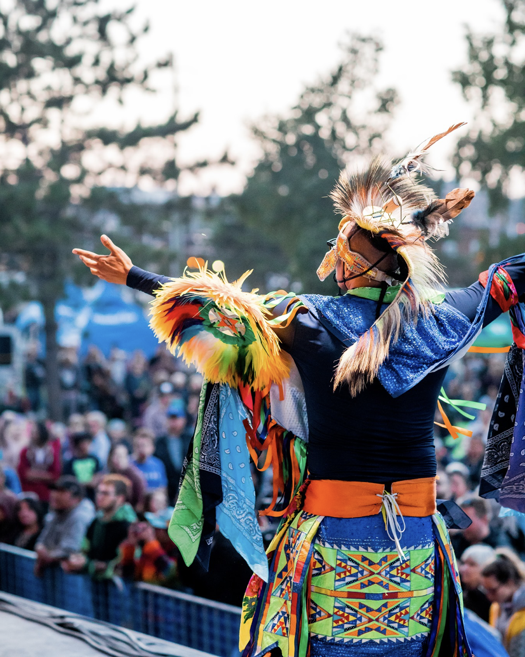 An indigenous man in traditional celebratory dress stands on a stage facing a crowd below with open arms. Green trees and a sunset are in the background.