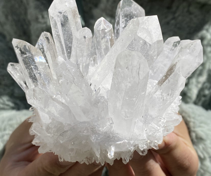 A large cluster of quartz crystals held by two hands in front of a grey stone background