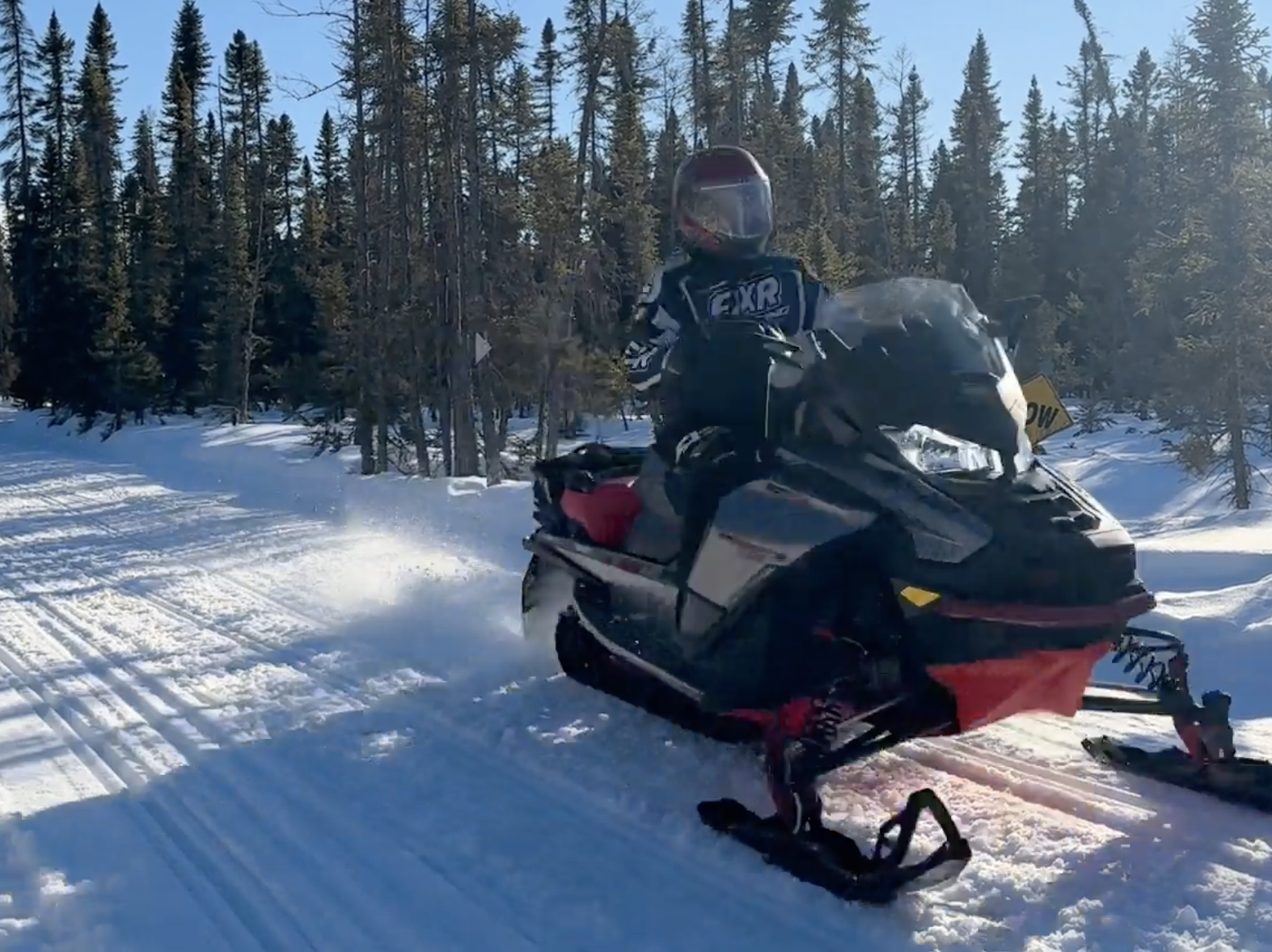 a snowmobiler riding down a snowy forested trail on a sunny day