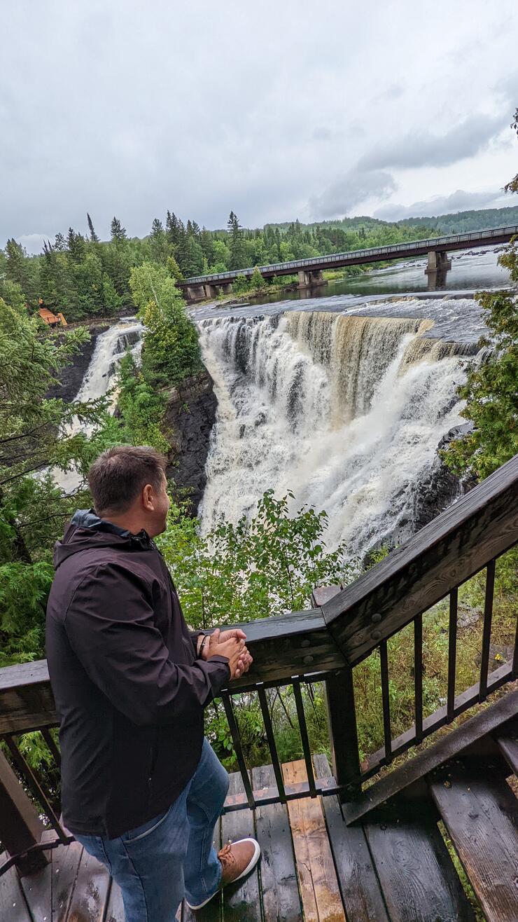 a tourist stands on a footbridge and looks out over Kakabeka Falls; a tall waterfall surrounded by lush green trees