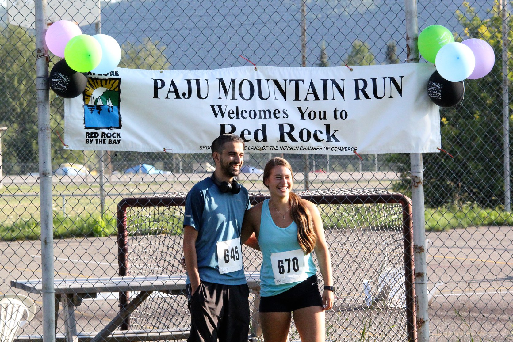 Two runners with arms around each other smile in front of a banner that reads "Paju Mountain Run Welcomes You to Red Rock"