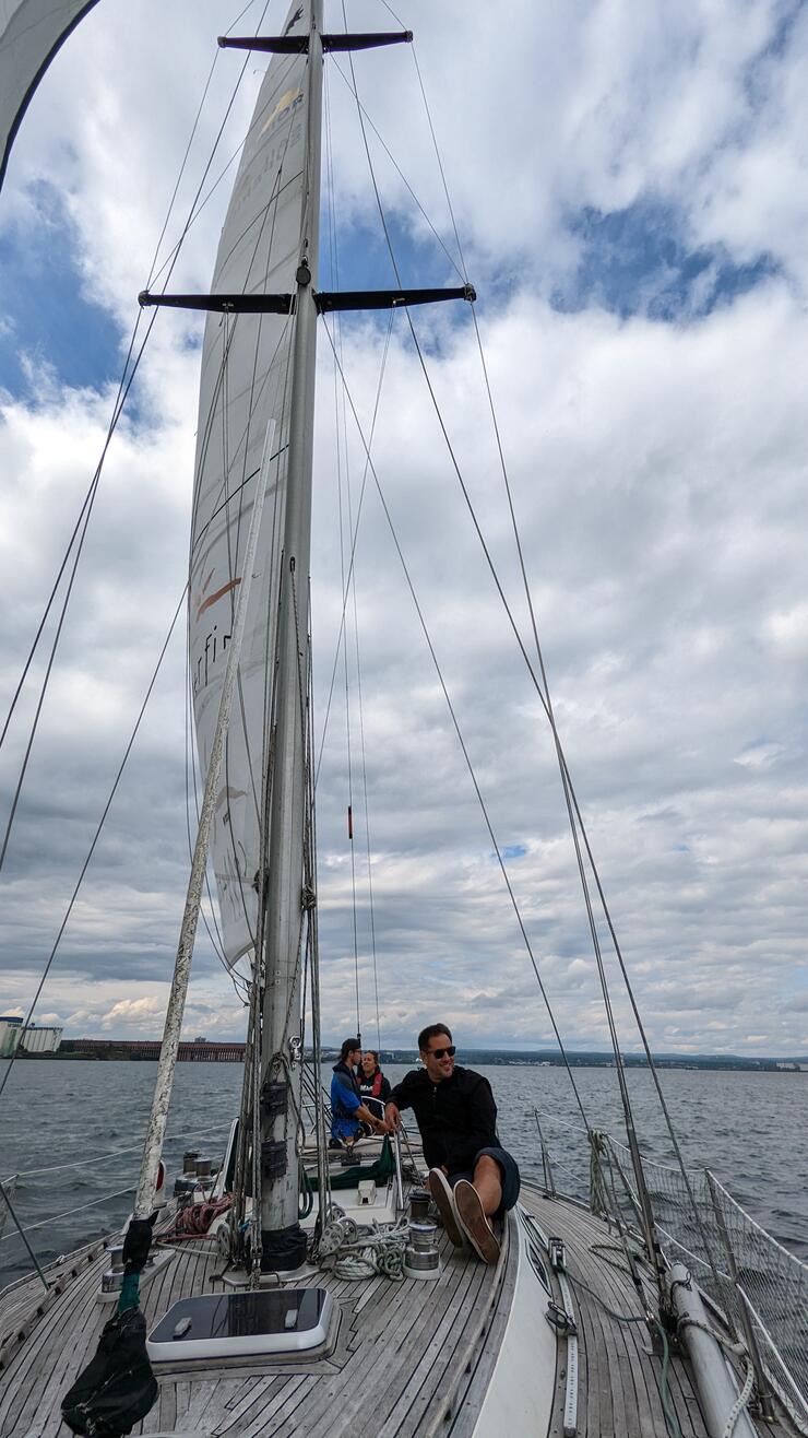 a man smiling, sitting on the deck of a large sailboat on open water.