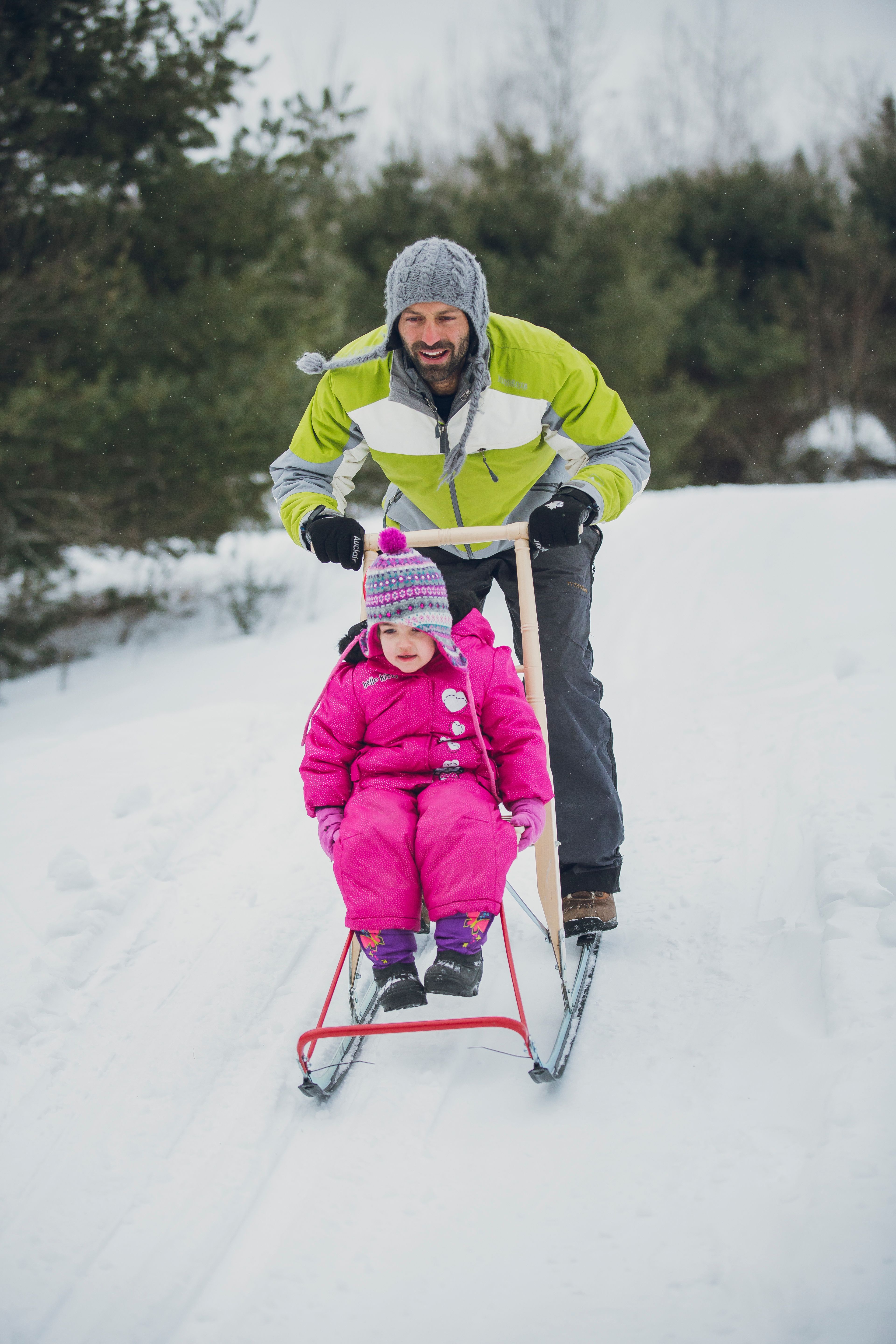 A  smiling man pushes a child on a kicksled through the snow. There is conifer forest in the background and snow is falling.