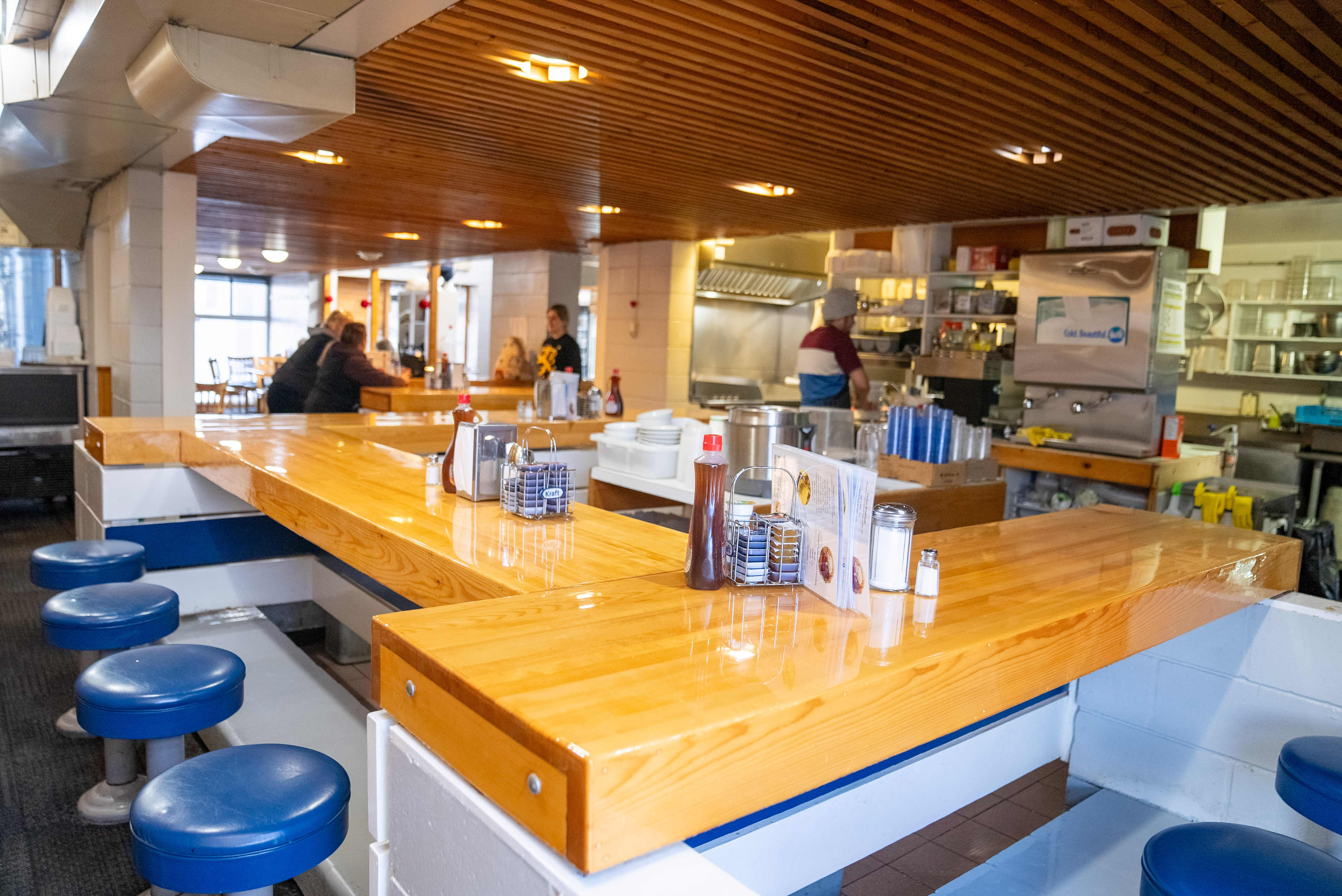 the cafe at Kanga's Sauna, Thunder Bay; a well-lit and shiny, inviting wooden breakfast bar surrounded by blue stools and adorned with condiment jars, with customers ordering at a chef's counter in the background.