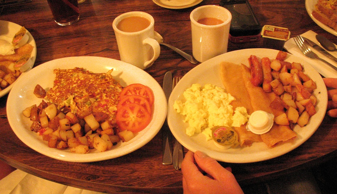 two plates of finnish pancakes with sides of hashbrowns, eggs and tomatoes on a table with cups of coffee.