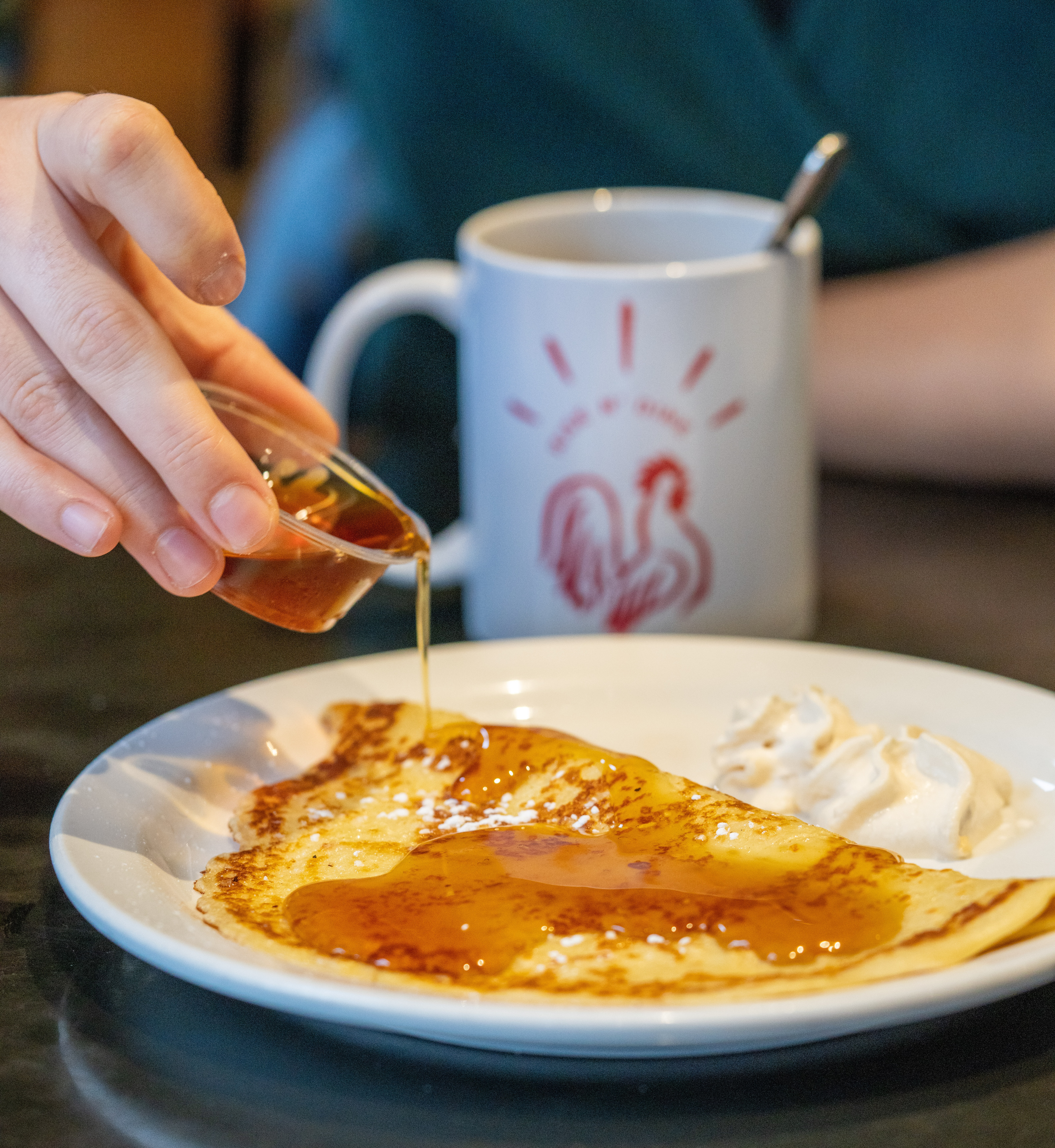 a finnish pancake on a plate, being drizzled with maple syrup. In the background there is a coffee mug with a red rooster logo on it.