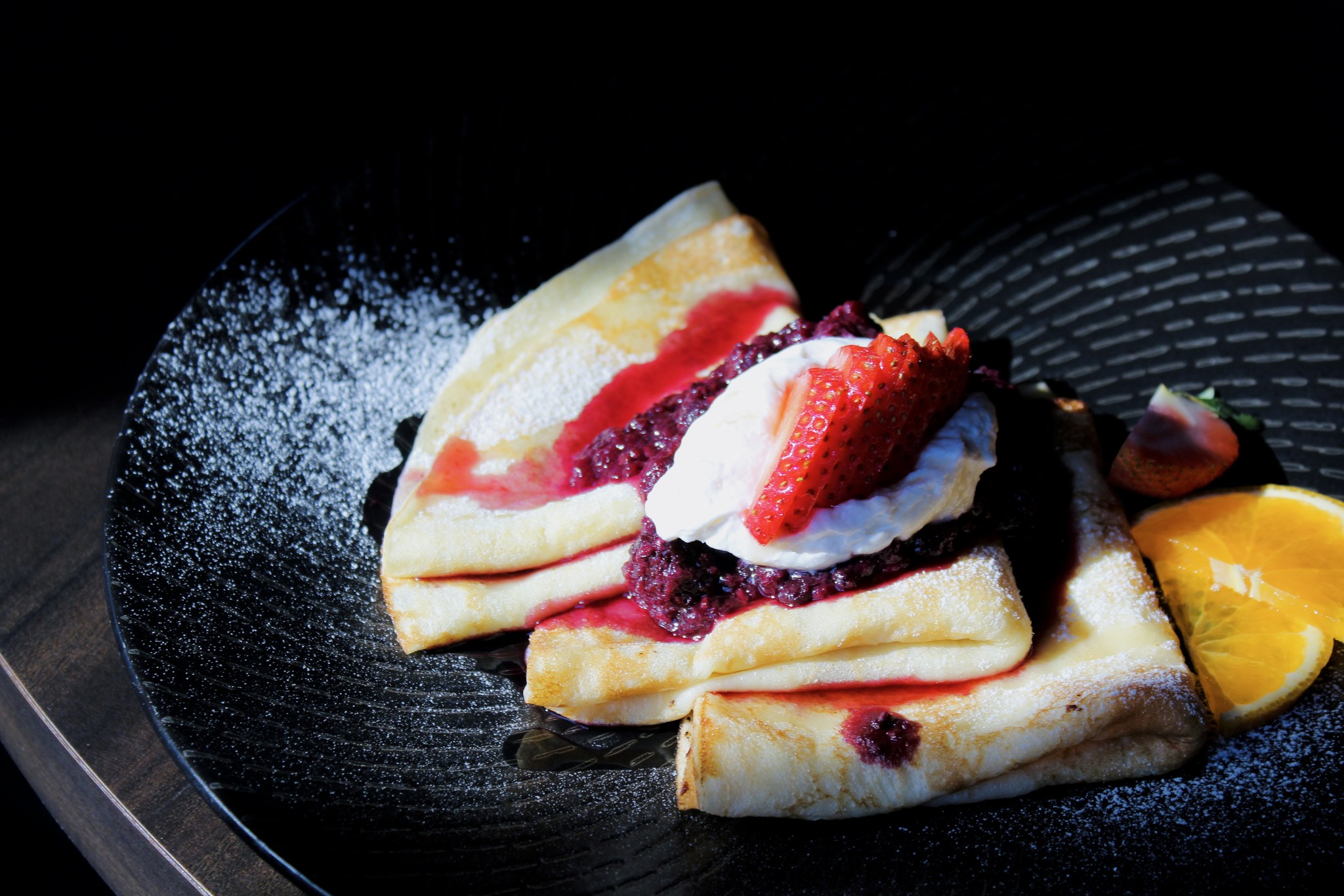 Skal Finnish pancakes with 3-berry compote, folded in thirds and laid out on a black plate and dusted with powdered sugar.