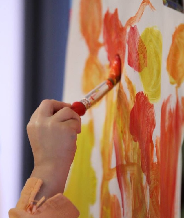 a child's hand holding a paintbrush, painting dots of yellow and orange on a large paper.