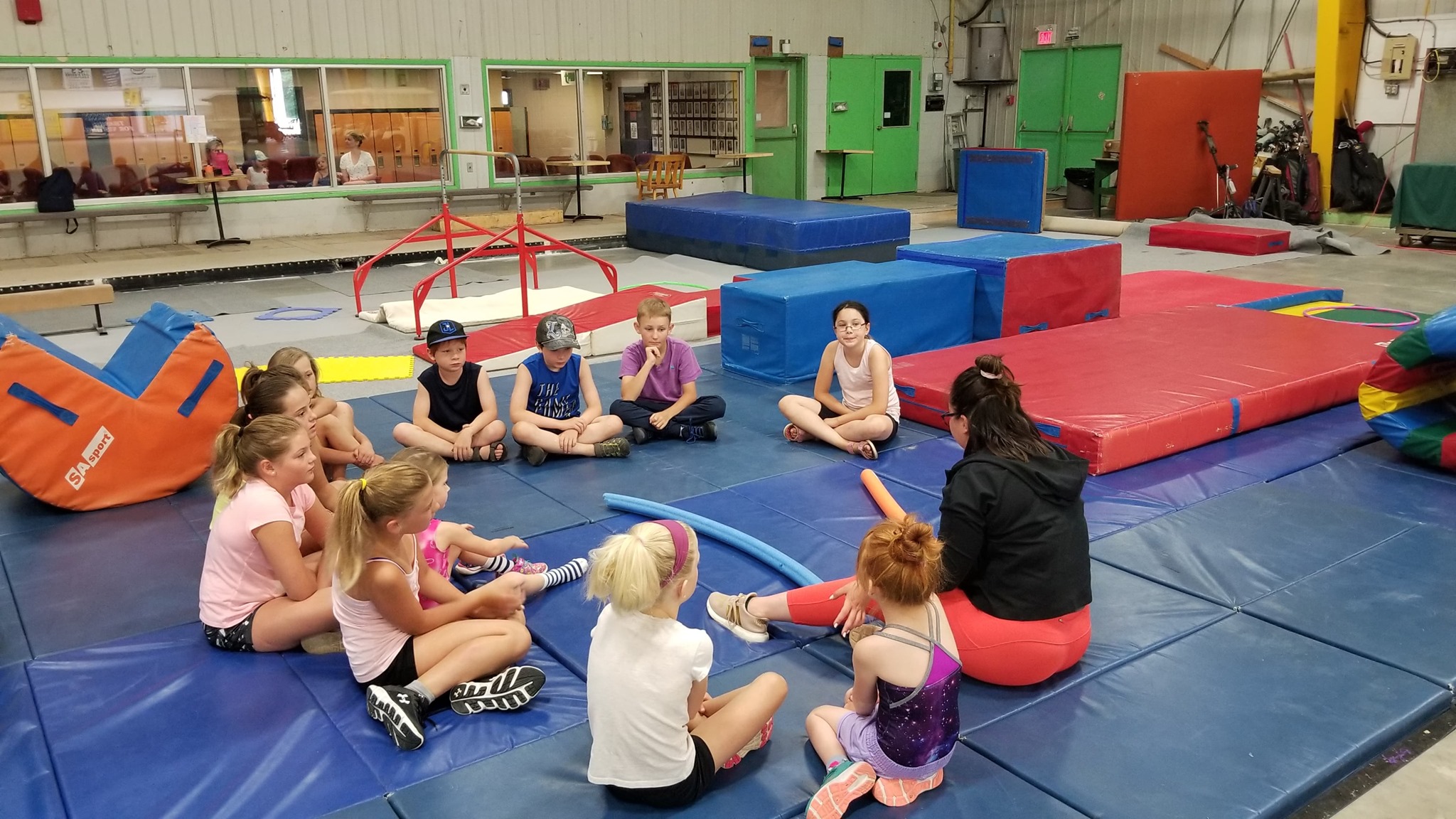 children and coaches sit in a circle in a gym full of colourful mats and climbing apparatuses.