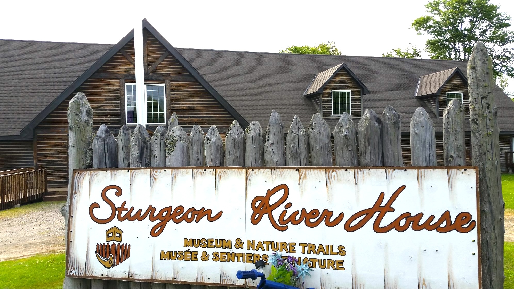 The Sturgeon River House museum; a rough wood building surrounded by green grass and trees with an aged but well-kept wooden gate and a large sign that reads "Sturgeon River House".