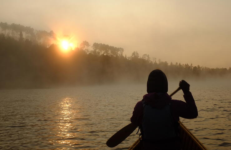 Canoeist paddling into sunrise on misty lake.