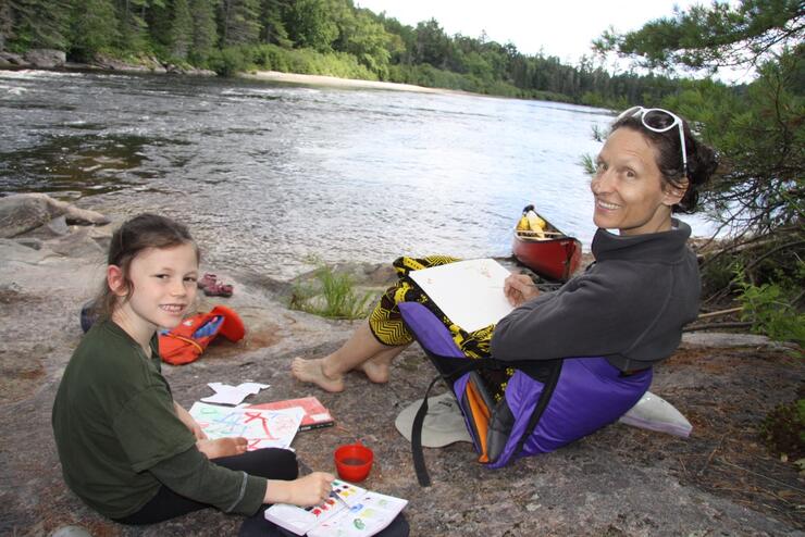 Mother and daughter sitting on rocks painting beside a river