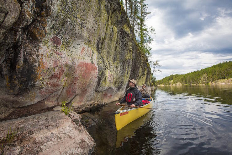 Tow men in a canoe paddling along a rock face with pictographs.