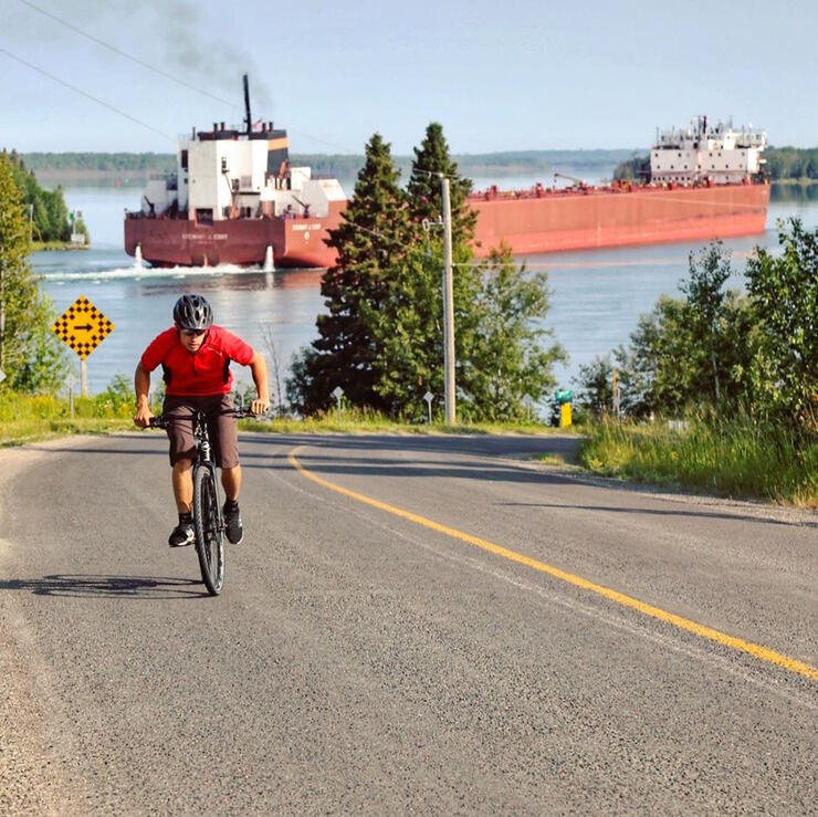 Male cyclist pedalling up a hill with a boat in background.