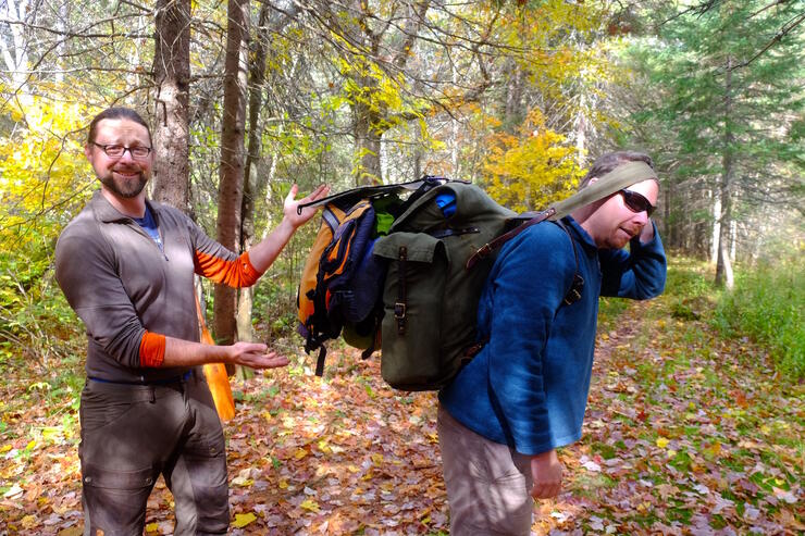 One man with heavy backpack and one man pointing at him.