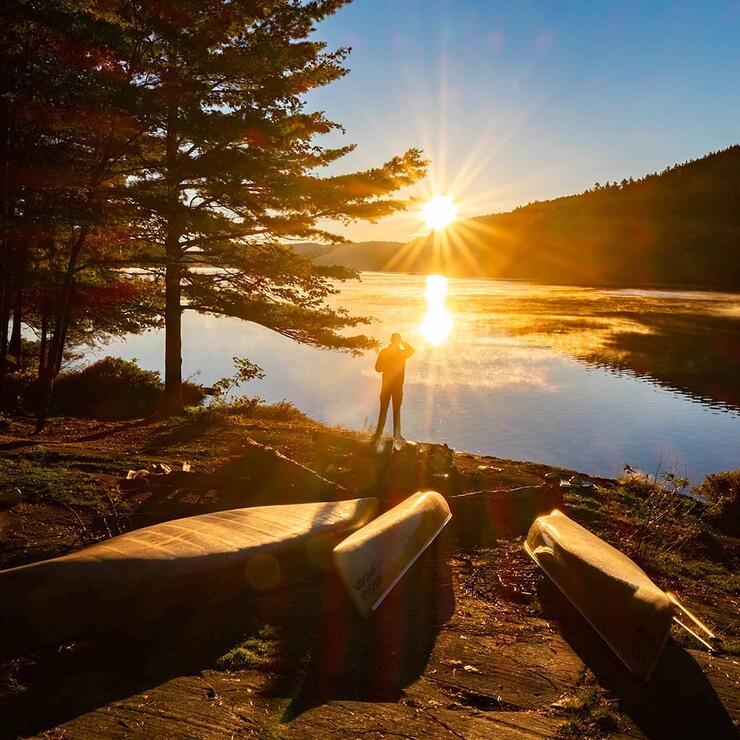 Beautiful sun burst over calm water. Man standing with canoes on beach.
