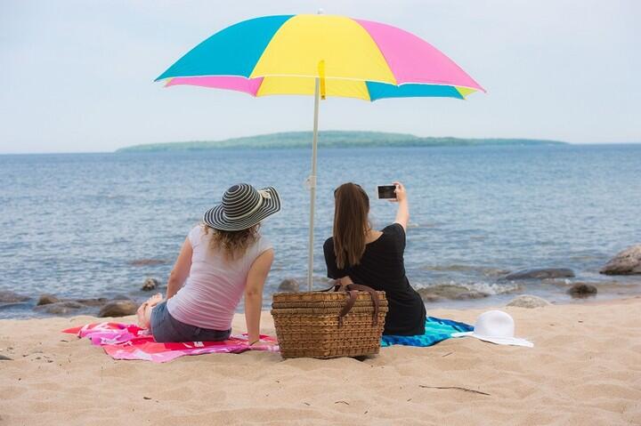 two women sit under umbrella on a beach and take photos of Giant’s Tomb Island from Awenda Park