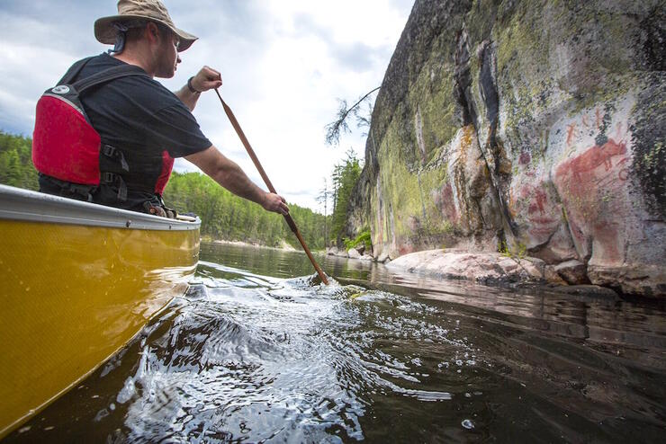 Man in a canoe paddling beside a rock face with pictographs