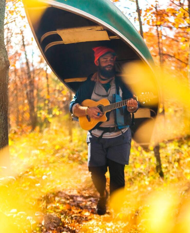 Man strumming on a guitar while portaging a green canoe on his head. 