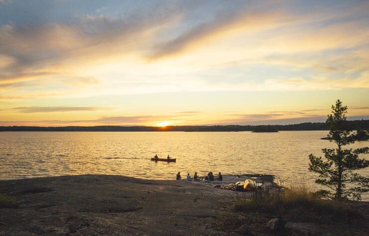 Group relaxing on smooth rocky shore at sunset. Canoe paddling by.