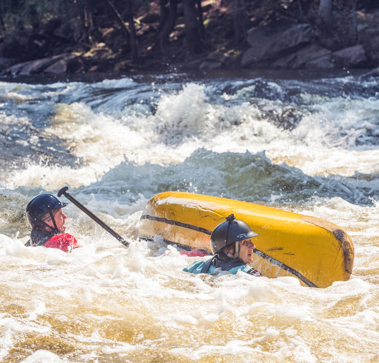 Two paddlers in whitewater beside overturned yellow canoe
