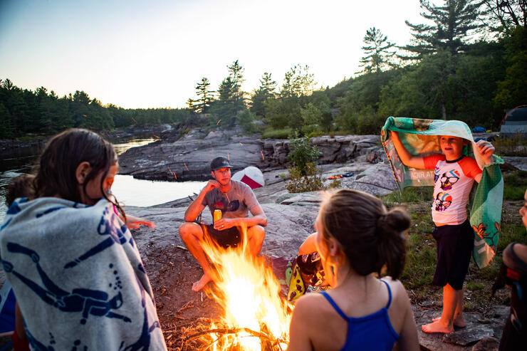Kids and adults gathered around a campfire by a river.