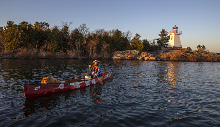 Man and a dog in a canoe paddling by a lighthouse.
