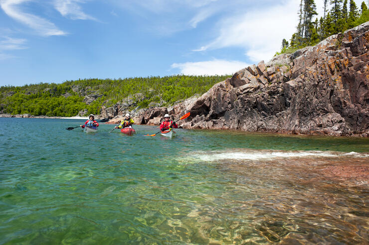 Three kayakers paddling in turquoise waters along a remote rocky shoreline