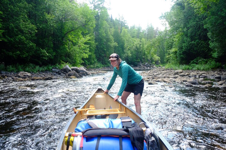 A woman standing in shallow rapids at bow of packed canoe.