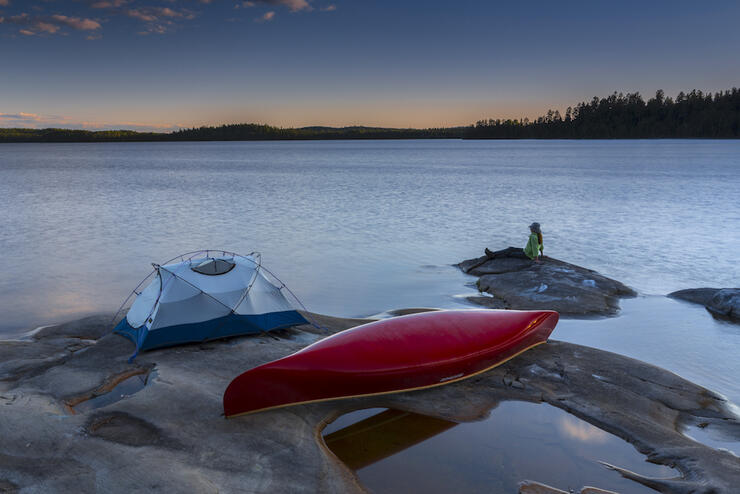Woman lounging on a smooth rock at lakeshore, beside a tent and canoe, at sunset