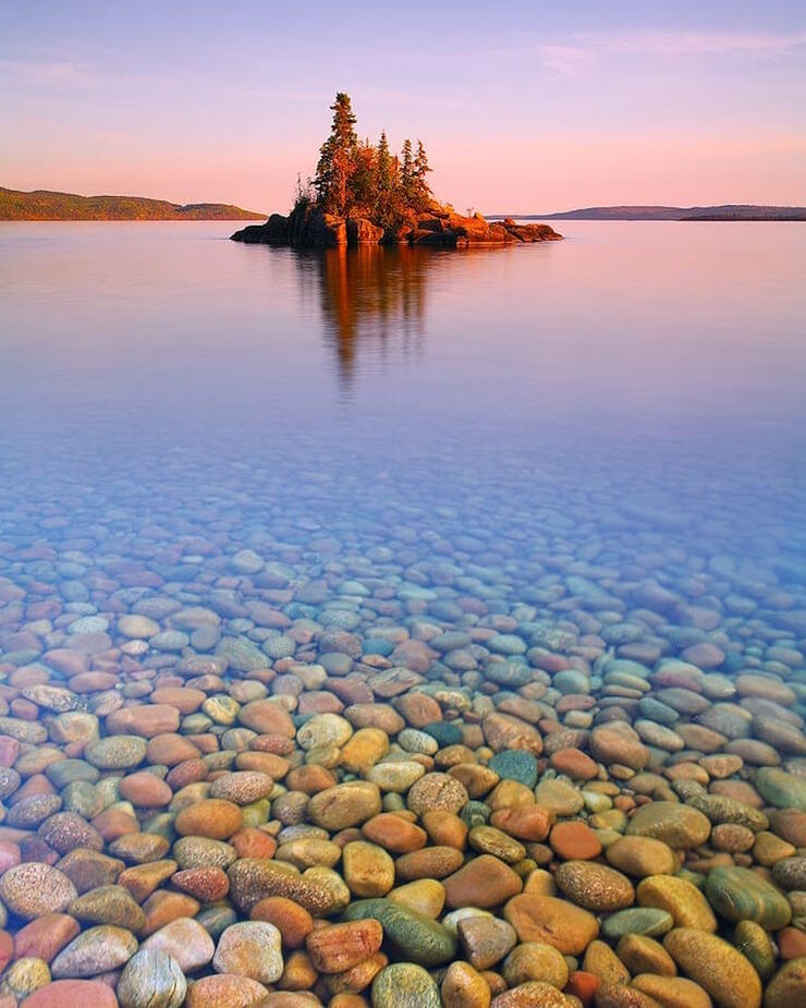 A distant island glowing in the pink hue of sunrise with rounded rocks in foreground. 