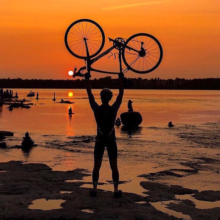 A man holding a bicycle upside down over his head with orange sunset in background.