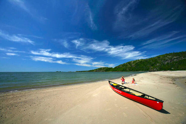 Woman and young girl sitting on an empty sand beach with red canoe in foreground