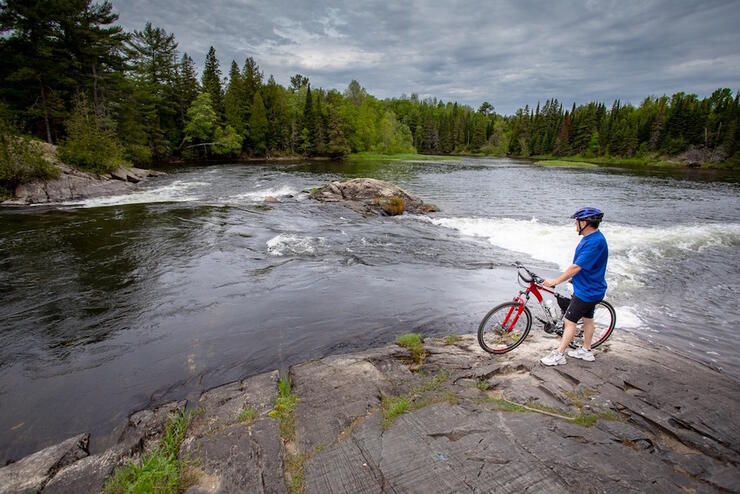 Man standing beside a bicycle on a rocky shore overlooking a rapid.