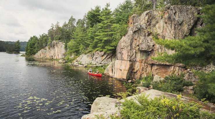 Two people in a red canoe paddling along shore of lake beside rock cliff.