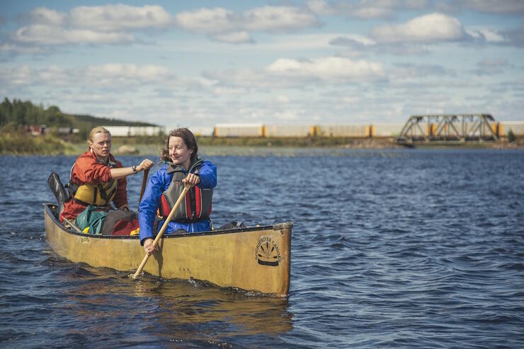 Man and woman paddling on a lake, with train in background.