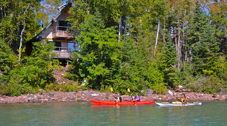 Kayakers paddling by a log cabin.