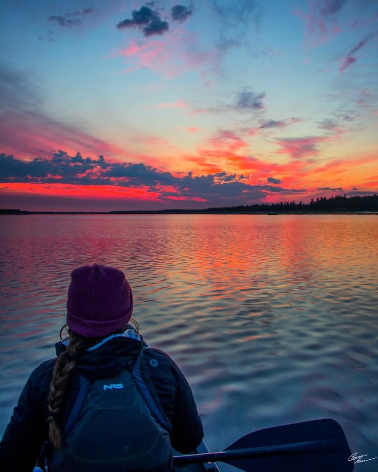 Person in the bow of a canoe, paddling into sunset.