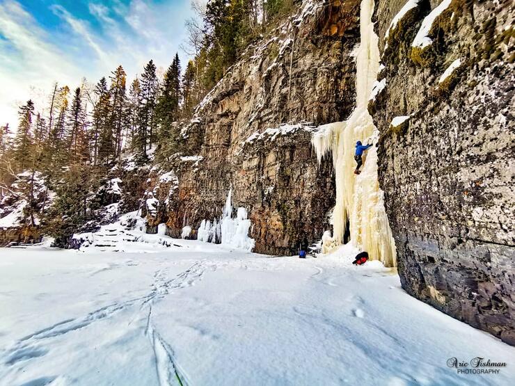 A climber ascending an ice covered rock face beside a snow covered lake.