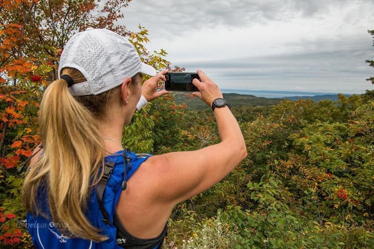 Woman in baseball cap with blond ponytail taking a picture of view with phone.