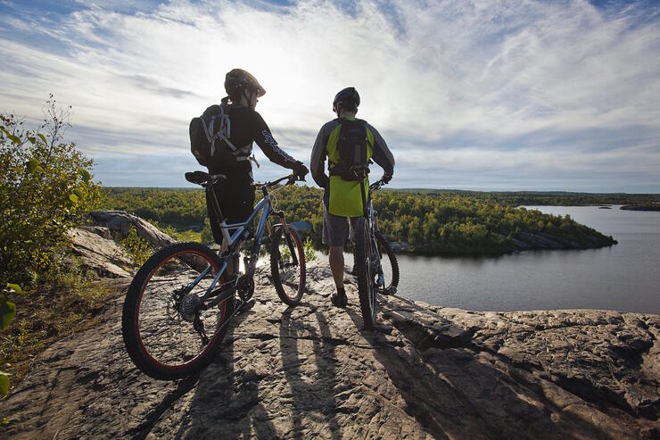 Two cyclists standing with bikes on a rocky lookout overlooking a lake.