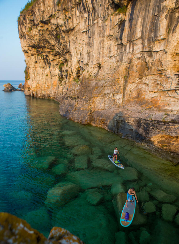 Two people stand-up paddle boarding along side tall cliffs