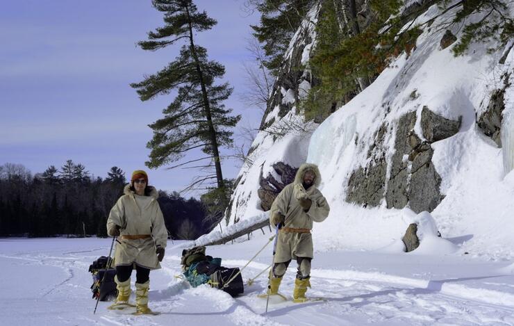 Man and woman in traditional winter gear pulling toboggans on snow-covered lake beside a rock face