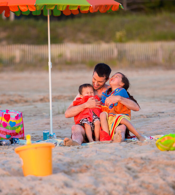 Man sitting on a beach under an umbrella hugging two laughing kids. 
