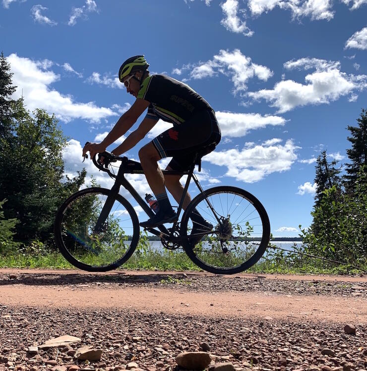 Man on a mountain bike riding on a gravel road with a lake and clouds in background.