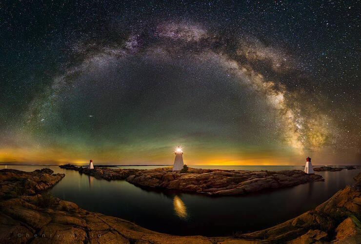 Star-filled sky with 3 lights houses on a smooth rocky shoreline.