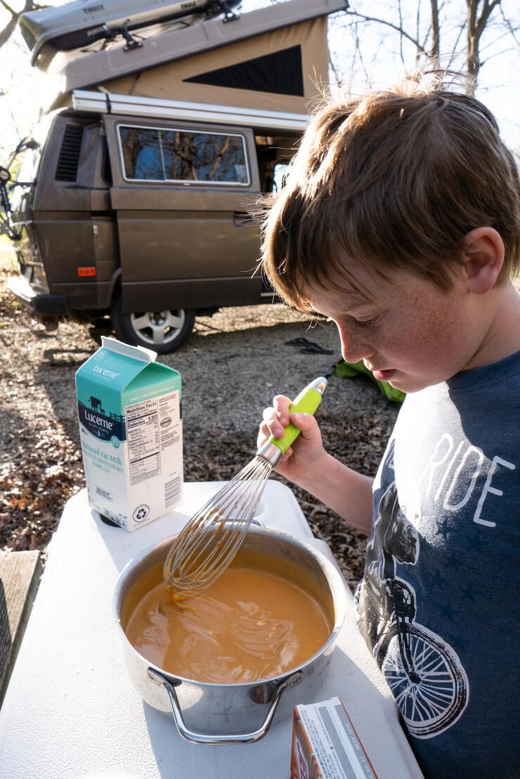 A young boy stirring a pot of pudding with a camper van parked in background.